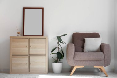 Interior of light living room with wooden drawers, blank frame and armchair