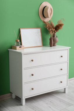 Chest of drawers with blank frame, vases, reed diffuser and box near green wall
