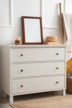White chest of drawers with blank frame, books and decor near light wall