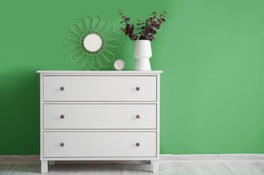 Vase with eucalyptus, alarm clock and mirror on chest of drawers near green wall