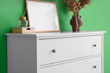 Chest of drawers with blank frame, vases, reed diffuser and box near green wall