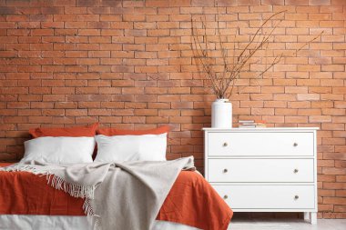 Vase with tree branches, books on chest of drawers and bed near brick wall