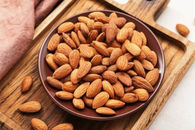 Wooden board with plate of almond nuts on light background, closeup
