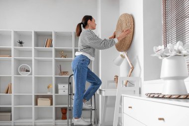 Young woman on stepladder hanging rattan pouf onto wall at home