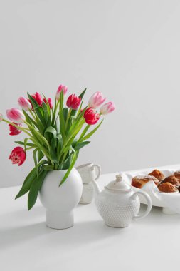 Vase with tulips, teapot and pastries on dining table near light wall