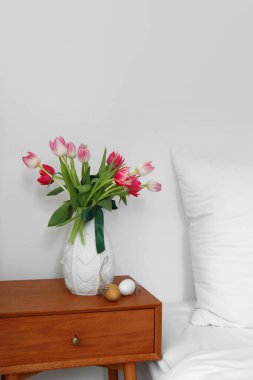 Vase with tulips and Easter eggs on table in light bedroom
