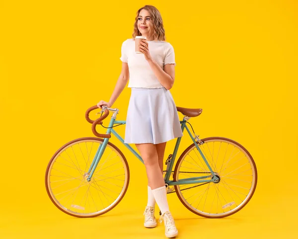 Happy young woman with bicycle and coffee on yellow background
