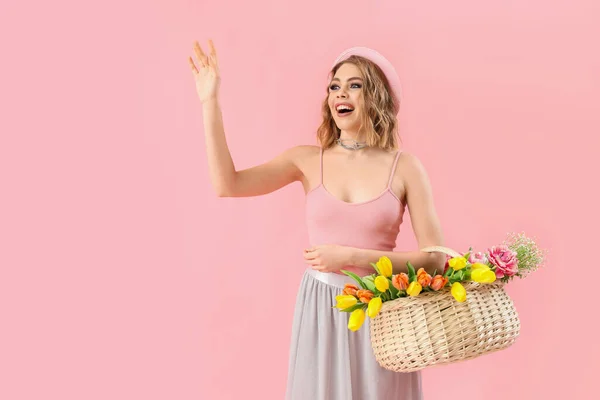 Beautiful happy young woman with basket of flowers waving hand on pink background