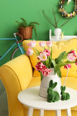 Vase with tulips and Easter rabbits on table near sofa in living room