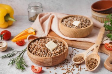 Wooden bowls of tasty buckwheat porridge with butter and vegetables on grey table