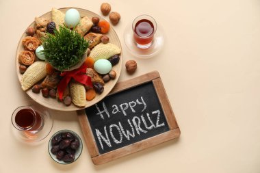 Chalkboard with text HAPPY NOWRUZ, treats, glass of tea and grass on beige background