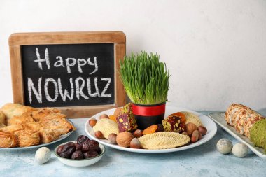 Chalkboard with text HAPPY NOWRUZ, treats, eggs and grass on table near white wall