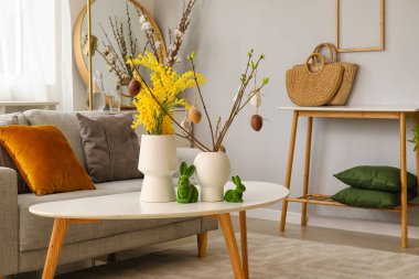 Vases with beautiful Easter decor and mimosa flowers on table in living room