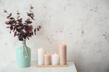 Burning candles and vase with eucalyptus on white table near grunge wall