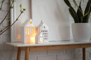 Glowing house candle holders and houseplant on table near white brick wall