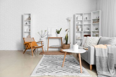 Interior of living room with armchair, sofa and house candle holders on table near white brick wall