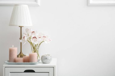Burning candles, vase with orchid and lamp on bedside table near white wall