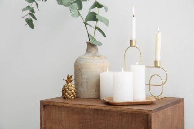 Candle holders with burning candles, houseplant and decorative golden pineapple on table near white wall