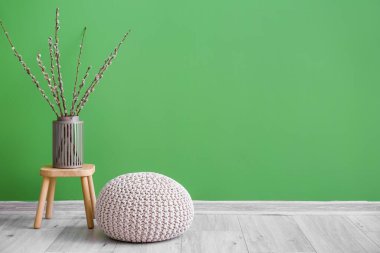 Vase with willow branches on stool and pouf near green wall