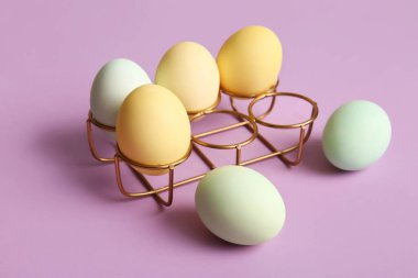 Holder with painted Easter eggs on lilac background