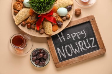 Chalkboard with text HAPPY NOWRUZ, treats and grass on beige background