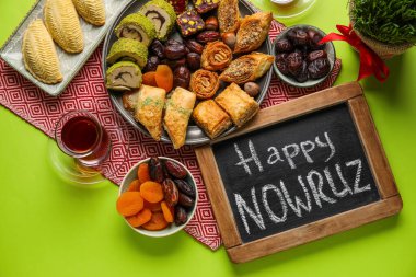 Chalkboard with text HAPPY NOWRUZ, treats, glass of tea and grass on green background