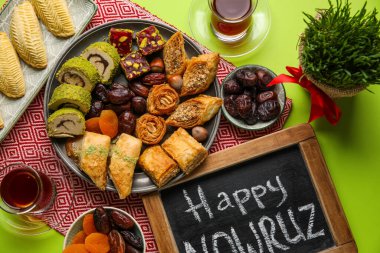 Chalkboard with text HAPPY NOWRUZ, treats, glass of tea and grass on green background
