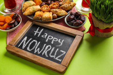 Chalkboard with text HAPPY NOWRUZ, treats and grass on green background, closeup