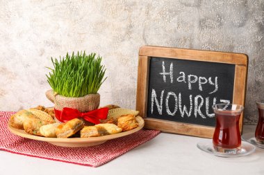 Chalkboard with text HAPPY NOWRUZ, treats, grass and glass of tea on table near grunge wall