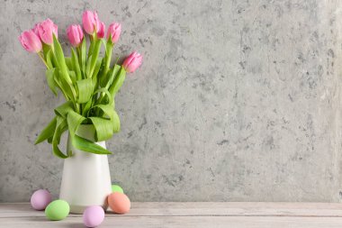 Vase with beautiful tulip flowers and painted Easter eggs on wooden table near light wall
