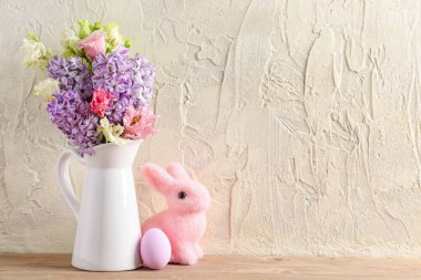 Jug with beautiful flowers, Easter egg and bunny on wooden table near light wall