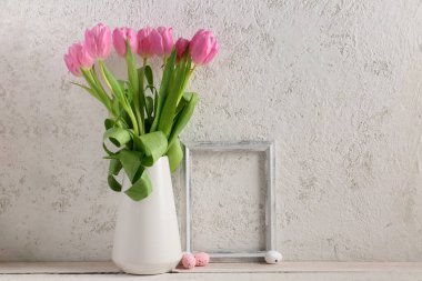 Vase with beautiful tulip flowers, Easter eggs and empty picture frame on table near light wall