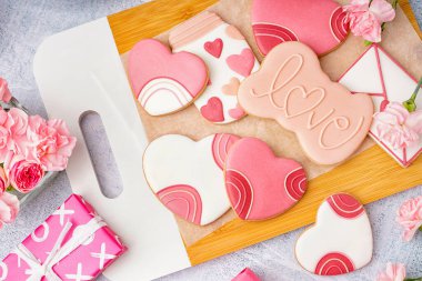 Wooden board with tasty cookies, gift and carnation flowers on light background, closeup. Valentine's Day celebration
