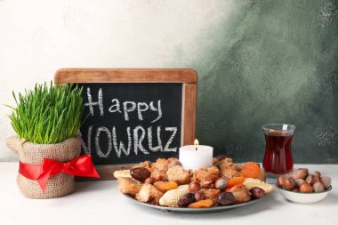 Chalkboard with text HAPPY NOWRUZ, grass, treats, candle and glass of tea on table near grunge wall