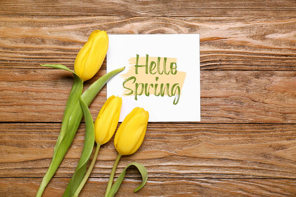 Card with text HELLO SPRING and yellow tulip flowers on wooden table