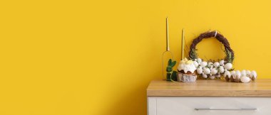 Easter cake and beautiful wreath with eggs on chest of drawers near yellow wall. Banner for design