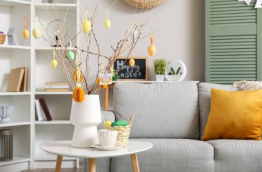 Vase with tree branches, Easter eggs and cup of coffee on table in living room