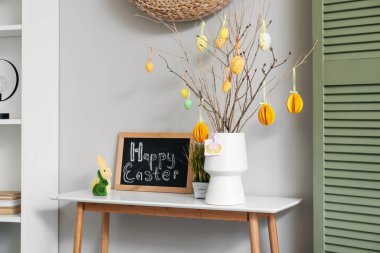 Chalkboard with text HAPPY EASTER, rabbits, grass and tree branches in vase on table near light wall