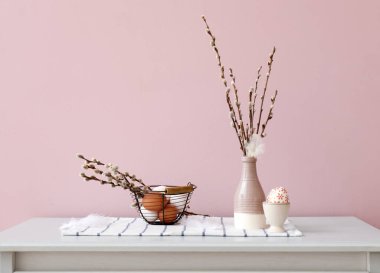 Vase with pussy willow branches, Easter eggs and feathers on drawer near pink wall