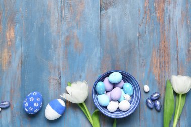 Composition with bowl of Easter eggs, candies and tulip flowers on color wooden background