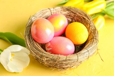 Nest with painted Easter eggs and tulip flowers on yellow background