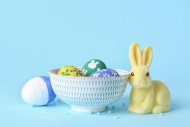 Bowl with painted Easter eggs and bunny on color background