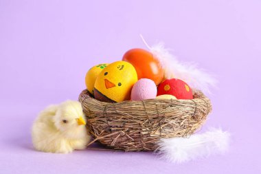 Nest with painted Easter eggs, feathers and toy chick on lilac background