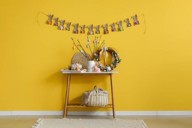 Tree branches decorated with Easter eggs, cakes and decor on table near yellow wall
