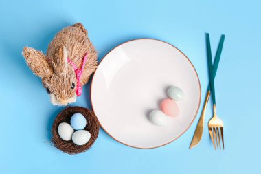 Table serving with Easter eggs, cutlery and bunny on blue background