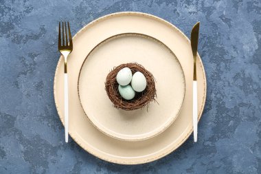 Table setting with cutlery and Easter eggs in nest on blue grunge background