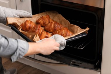 Male baker taking tray with croissants from oven in kitchen, closeup