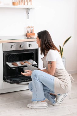 Young woman taking baking tray with cookies from electric oven in kitchen