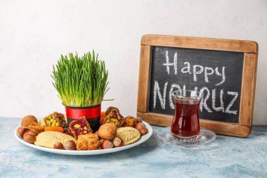 Chalkboard with text HAPPY NOWRUZ, treats, grass and glass of tea on table near white wall