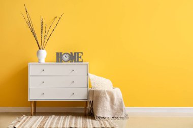 Vase with willow branches and clock on chest of drawers near yellow wall in room